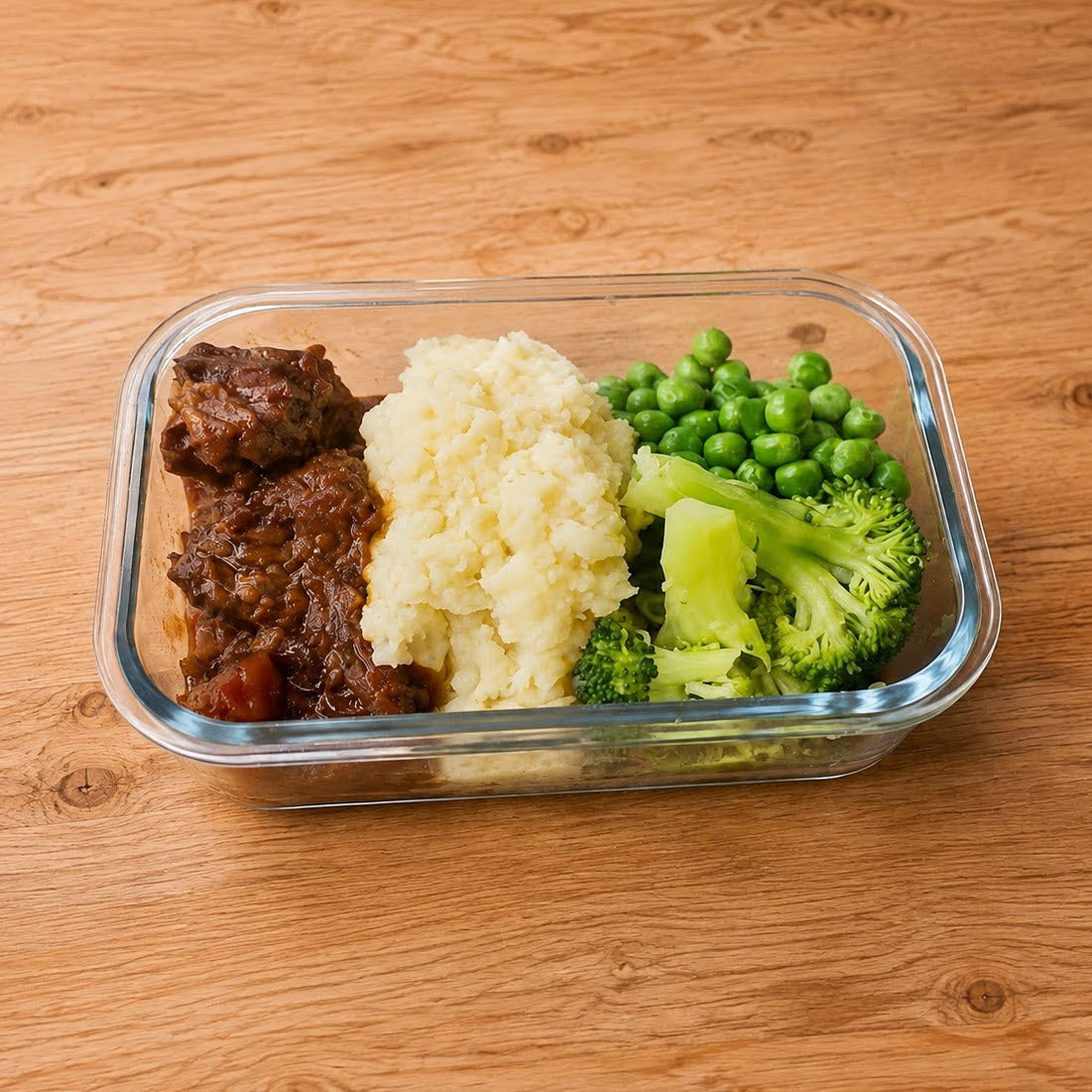 Glass meal container with slow-cooked beef, mashed potato, peas and broccoli on a timber table. 